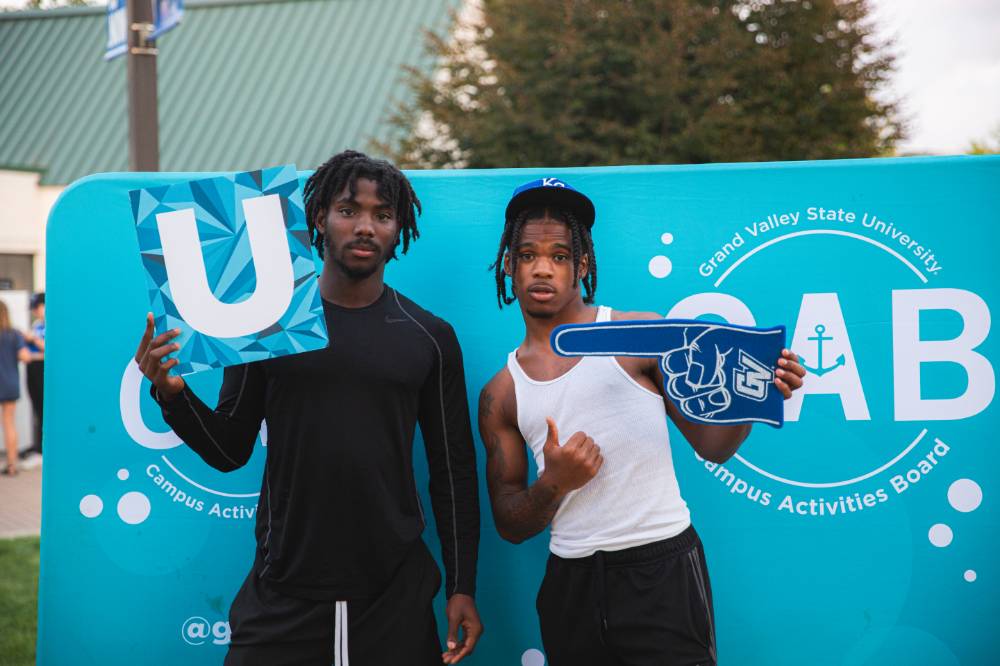 two students posing in front of CAB photo backdrop at Laker Kickoff photo booth and holding a U sign and foam finger